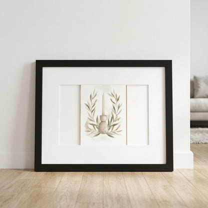 Framed wall art leaning against a white wall on a light wood floor, featuring a minimalist botanical print with olive branches and a small vase in a black frame with a wide white mat.