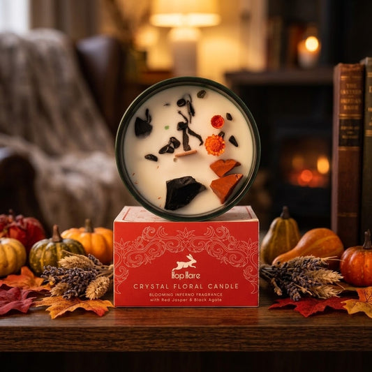 A wax candle in a round dark green tin sits on top of its red branded box on a wooden table, surrounded by fall decor including pumpkins, leaves, and dried flowers. The candle top shows embedded botanical pieces, and the box reads 'KapiKare Crystal Floral Candle'.