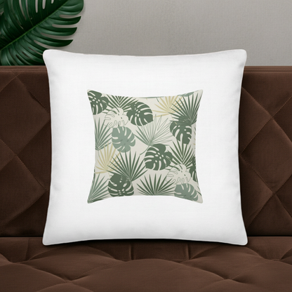 A square white throw pillow with a central printed panel featuring green and beige tropical leaf patterns, resting on a brown tufted sofa with a green leaf in the background.