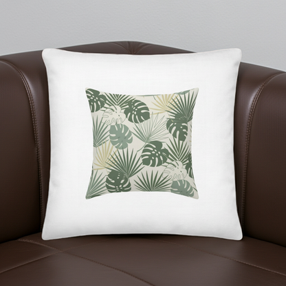 A square white throw pillow with a central green tropical leaf pattern, resting on a brown leather sofa against a light gray wall.