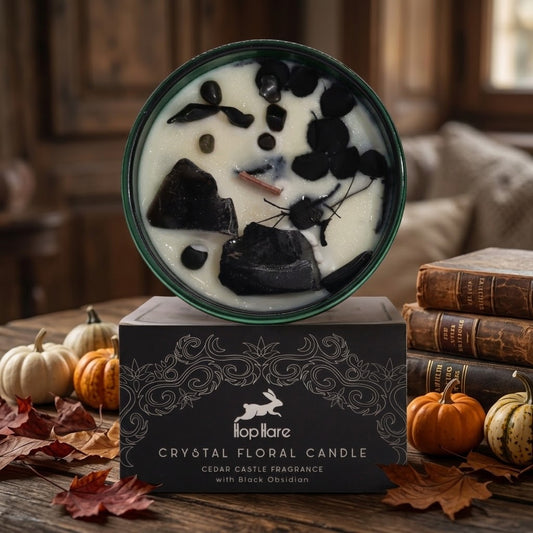 A round green-rimmed candle in a dark tin sits on top of its black branded box. The candle wax is creamy off-white with scattered black obsidian stones and a small dried botanical piece.