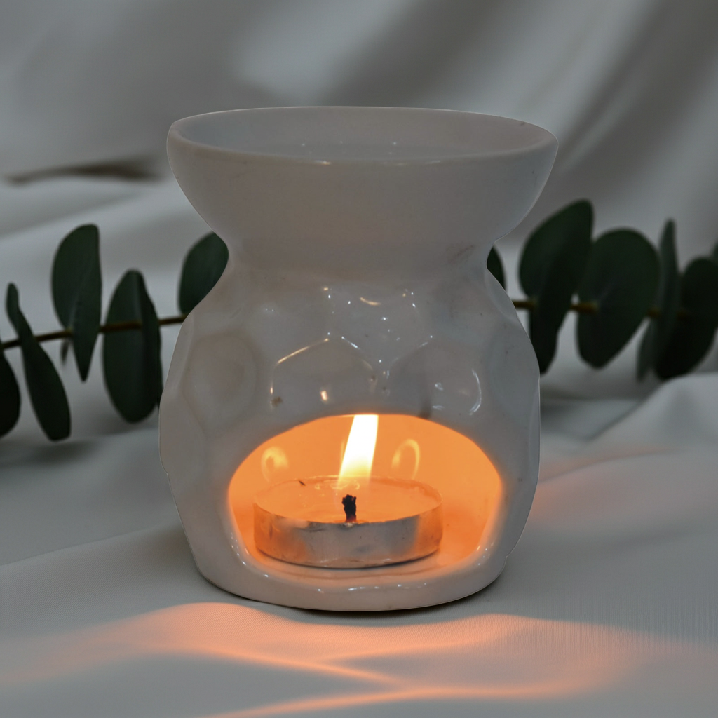 A white ceramic wax warmer holding a lit tealight candle, set against a draped white fabric backdrop with eucalyptus leaves in the background.
