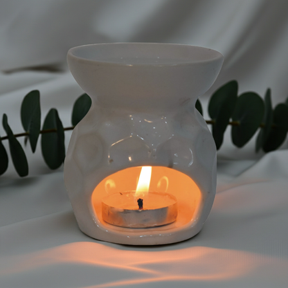 A white ceramic wax warmer holding a lit tealight candle, set against a draped white fabric backdrop with eucalyptus leaves in the background.