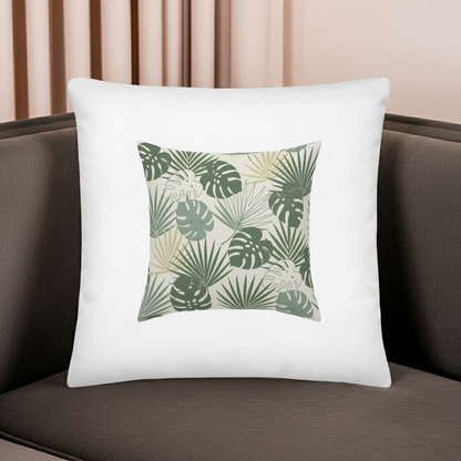 A square white throw pillow featuring a central tropical leaf pattern in shades of green and beige, resting on a dark brown sofa with a soft pink curtain background.