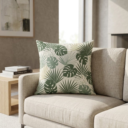 A square throw pillow with a green tropical monstera and palm leaf pattern on a cream background, styled on a light beige fabric sofa in a modern living room setting.