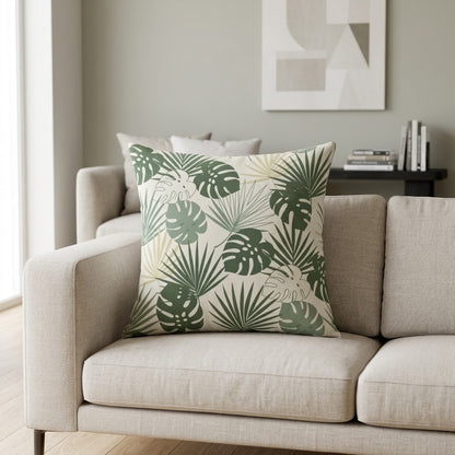A square throw pillow with a green tropical monstera and palm leaf pattern on a cream background, styled on a light beige sofa in a modern living room setting.